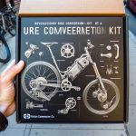 An HD image capturing the moment when a revolutionary bike conversion kit is being unveiled by Urban Commuter Co. The kit is presented as a practical solution for urban dwellers. It's known to transform traditional bikes into more efficient and environmentally friendly alternatives. The photo includes a detailed close-up of the high-tech components of the kit. The branding of Urban Commuter Co. is subtly displayed on the packaging and the parts of the kit.